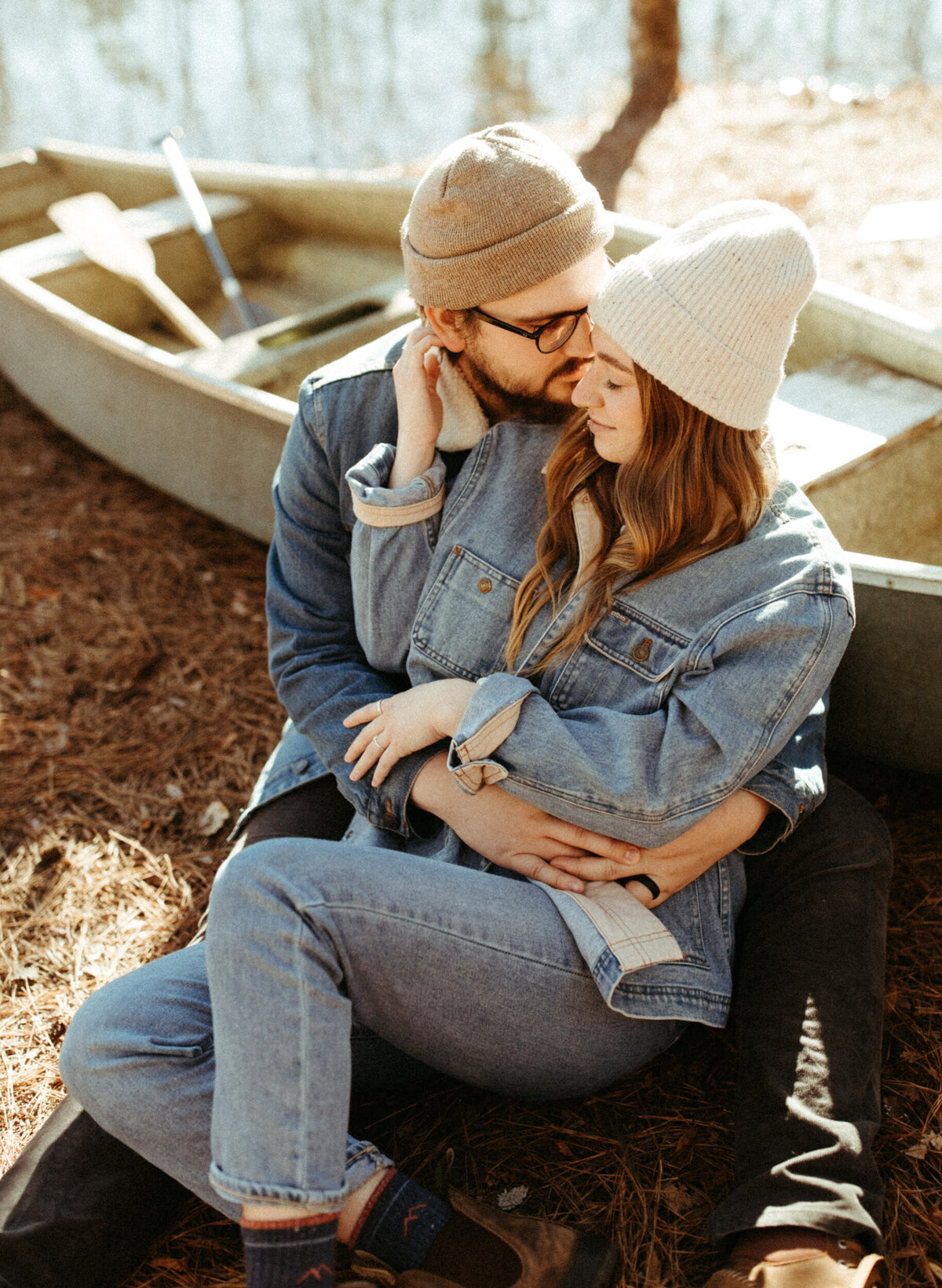 Couple in all denim outfits and beanies sitting down beside a canoe with a lake in the background
