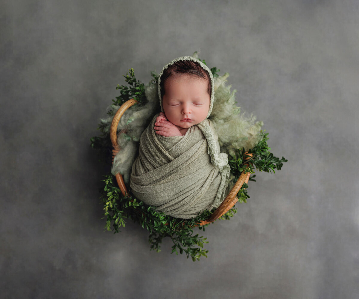 A newborn baby is peacefully sleeping, swaddled in a green blanket and lying in a round basket adorned with green foliage, set against a soft grey background.