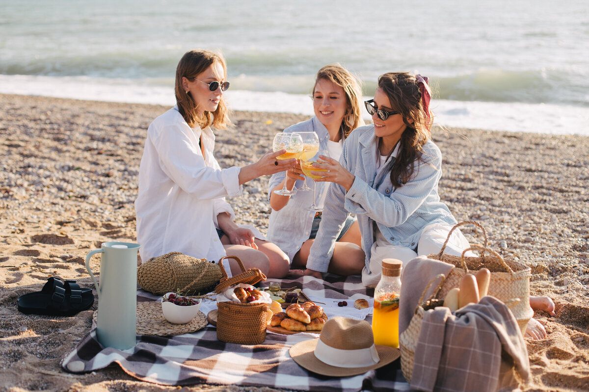 Three people sit on a picnic blanket at the beach, toasting with drinks. They are surrounded by baskets, pastries, and a bottle of juice. The ocean waves are visible in the background on a sunny day.