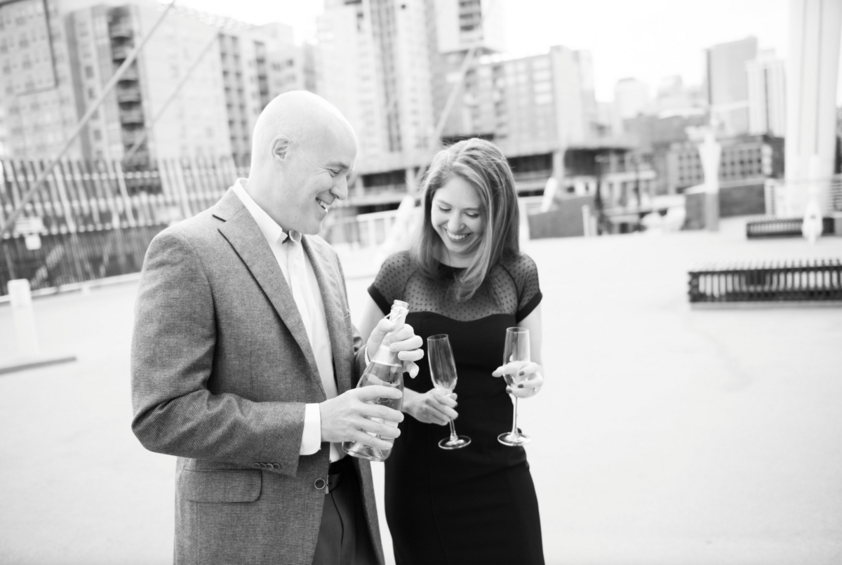 A man and woman laugh with one another as they share glasses of champagne during their downtown Denver engagement session.