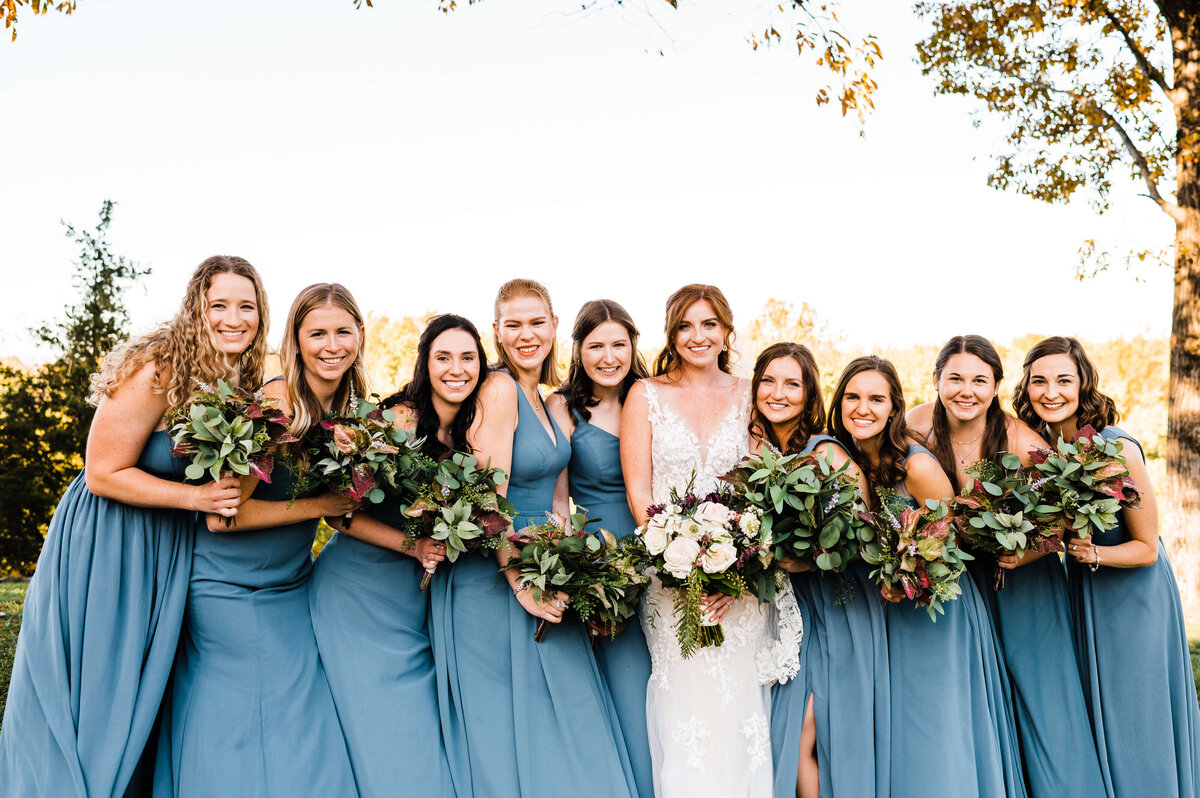 Bride in her wedding dress holding a bouquet of flowers surrounded on both sides by her bridesmaids who are in blue gowns holding bouquets at an outdoor Charlottesville wedding venues