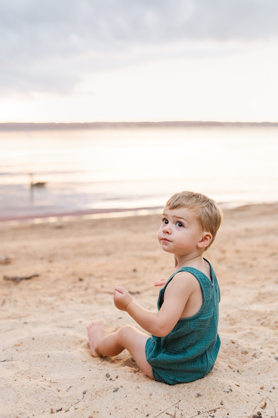 Little boy sits in sand at lakeside beach and stares up off camera