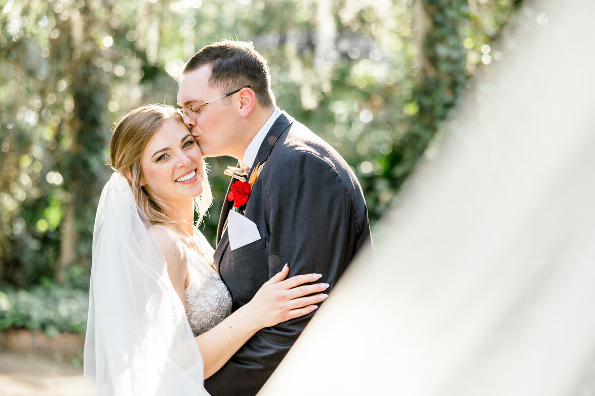 Groom kissing bride on temple as bride smiles at camera