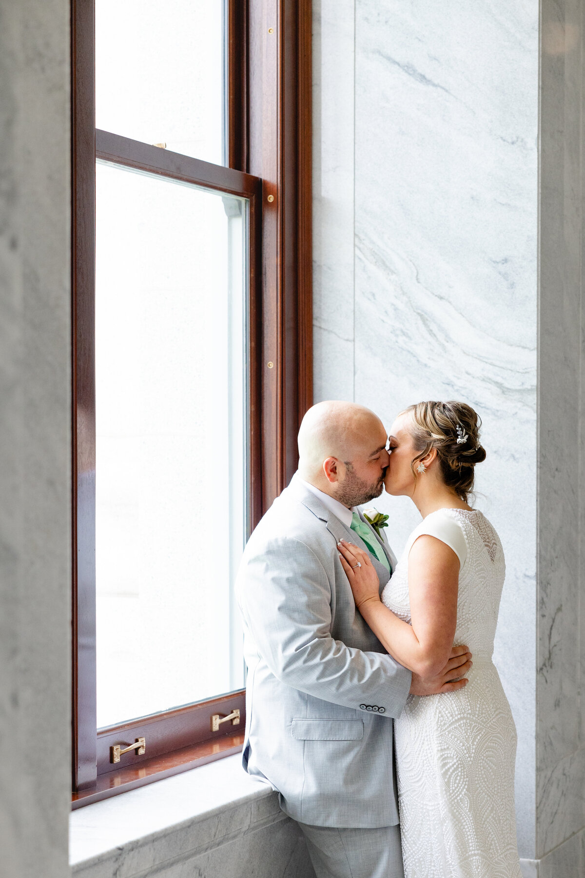 A newlywed couple shares a kiss by a large window at the Utah State Capitol, with natural light streaming in through the marble surroundings.
