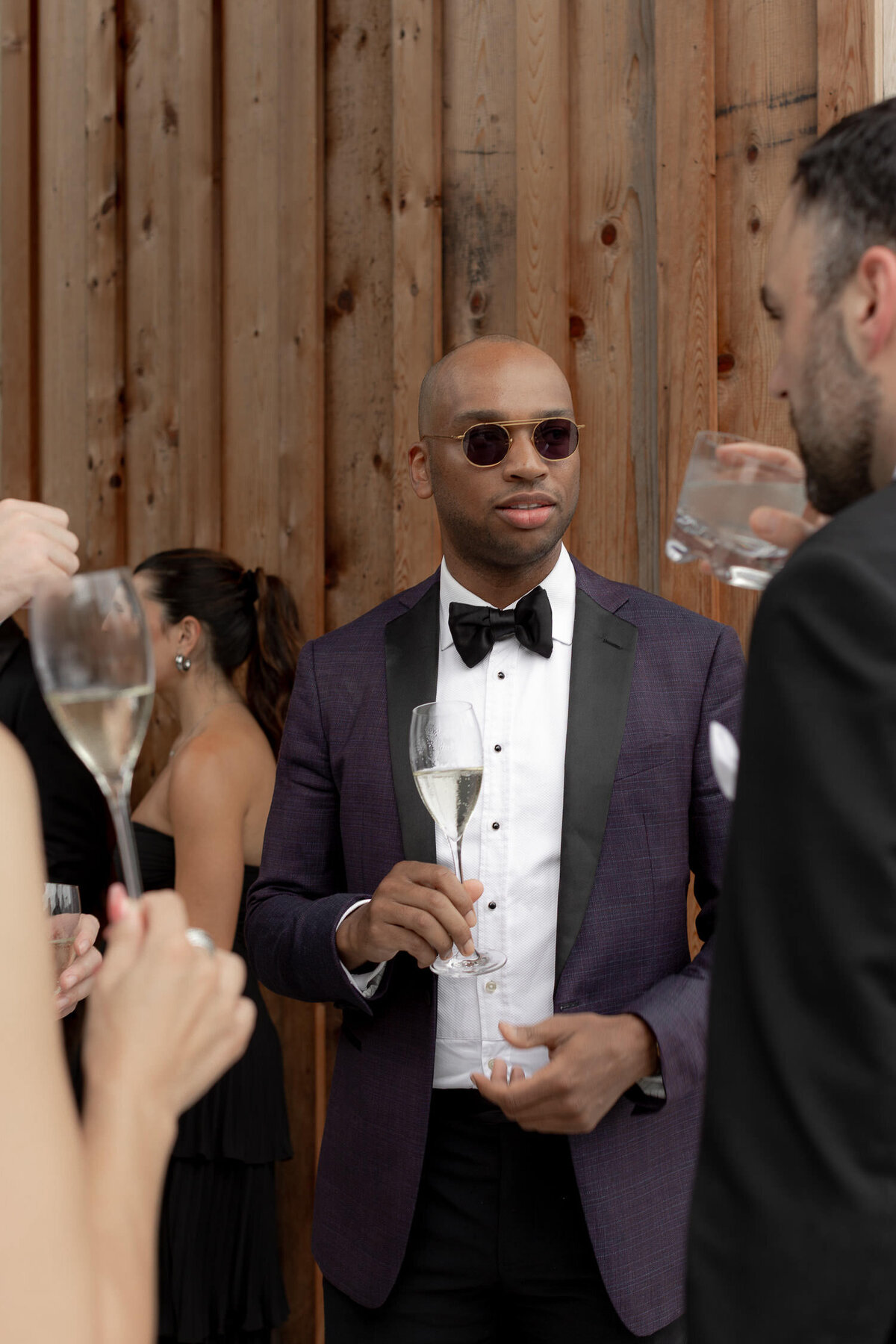 Wedding guest in purple tuxedo holding champagne glass and talks to guest. Wood panels in the background