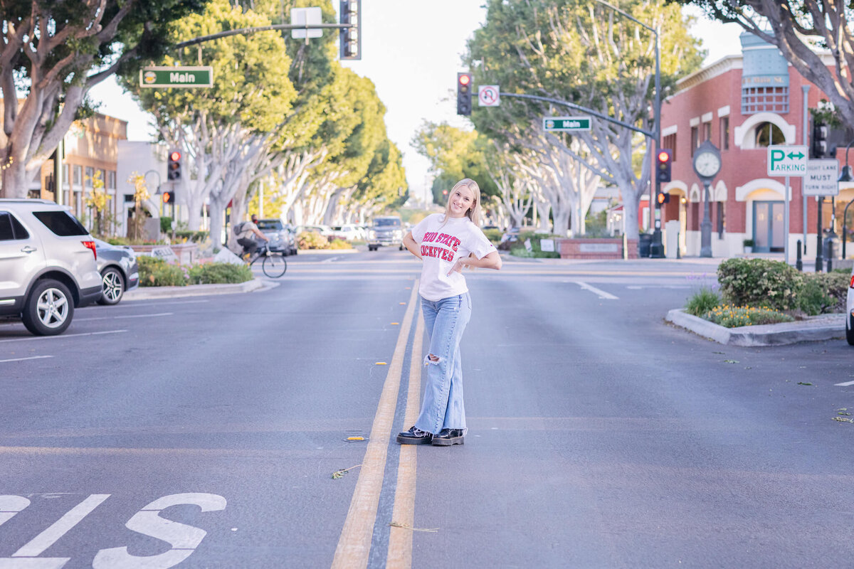 A person stands confidently in the middle of an empty street, wearing a white shirt and jeans. Trees line the sidewalk, and a traffic light and storefronts are visible in the background. The setting appears calm and sunny.