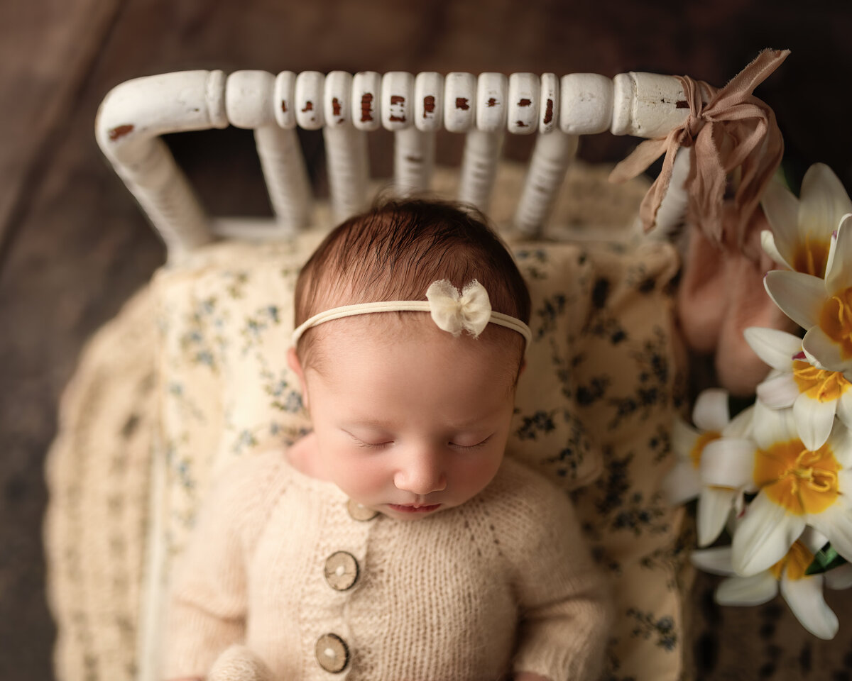 A beautifully styled newborn portrait taken at Meraki Photographic Creations in Paris, Ontario. Featuring a peacefully sleeping baby dressed in a hand-knit neutral sweater, resting on a vintage floral cushion with warm tones and rustic textures, surrounded by soft blooms.