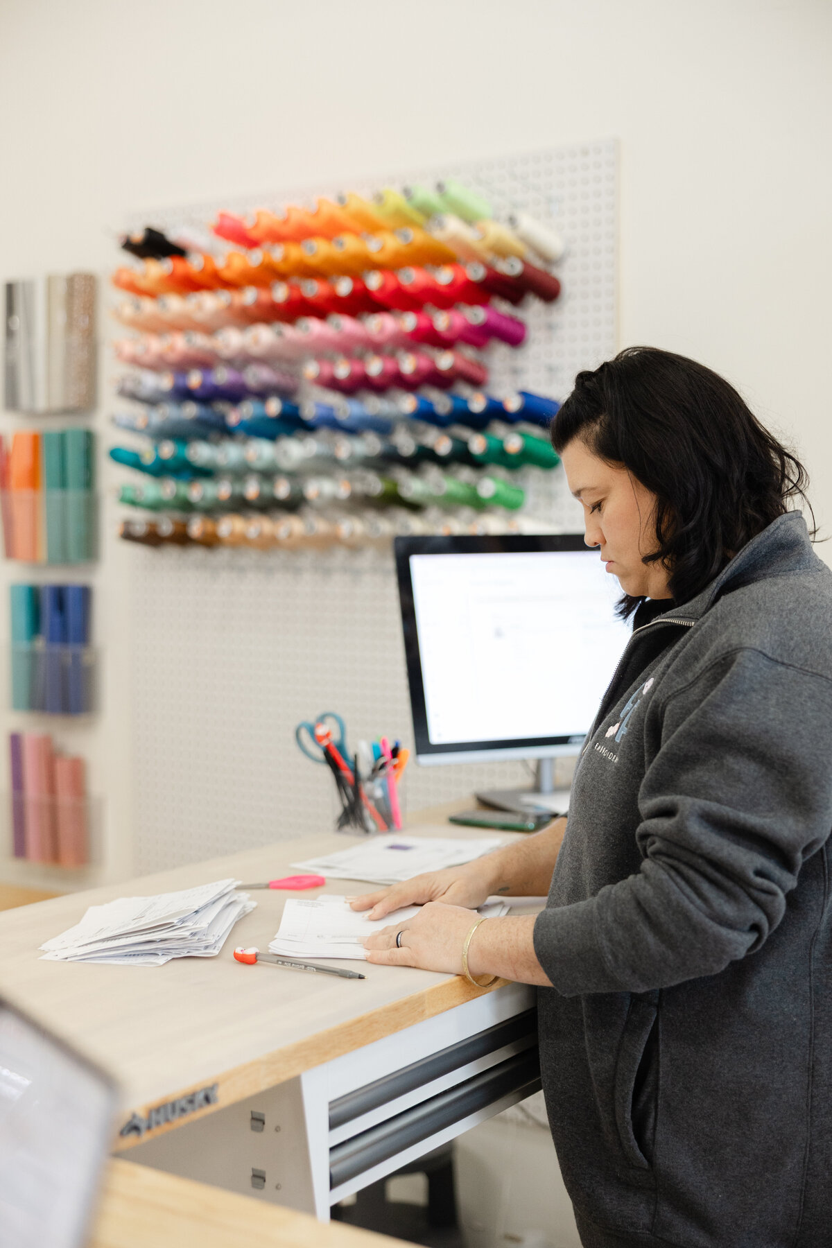Woman with dark hair working at embroidery machine beneath colorful thread display. Photograph by Yucaipa branding photographer Kaitlyn Dawn Photography.