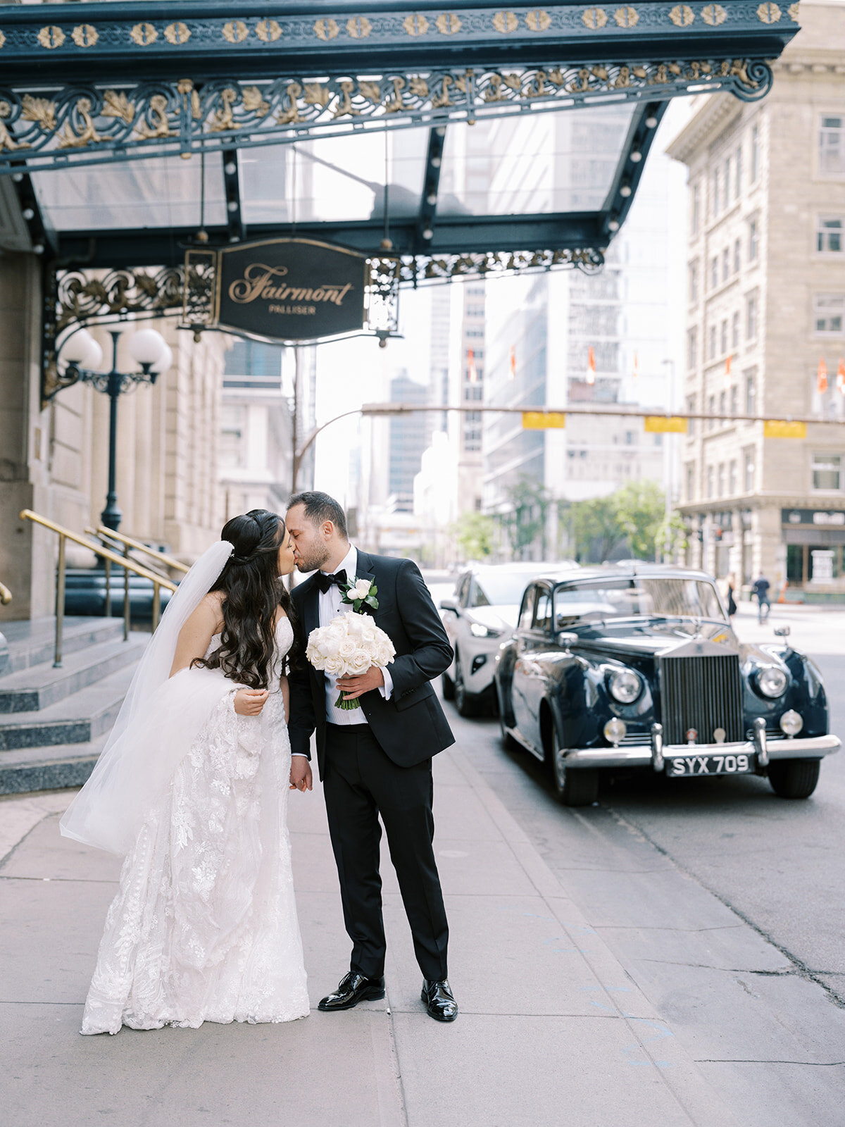 A bride and groom share a kiss on a city sidewalk in front of the Fairmont Palliser hotel, with a vintage car parked nearby. The bride, holding a bouquet, and the groom are elegantly dressed in formal wedding attire. It's an iconic scene for a classic Calgary wedding.