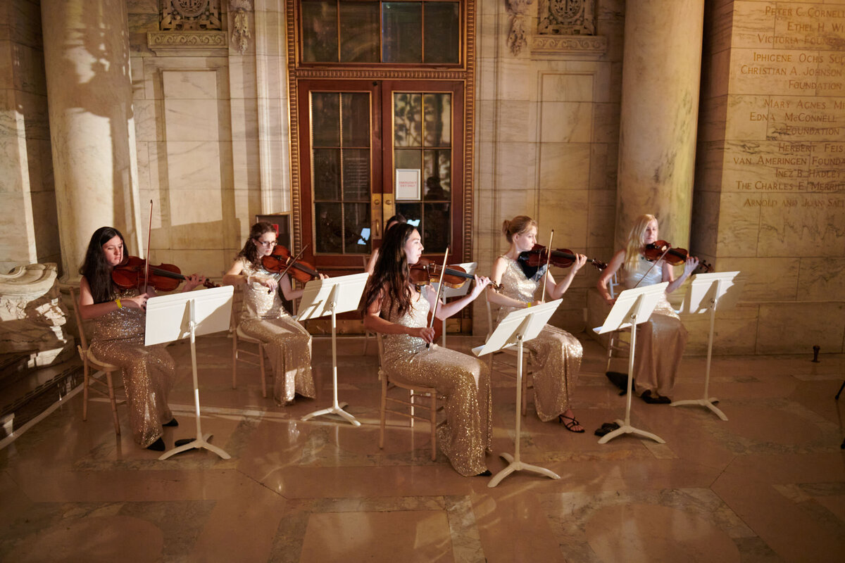 This captivating scene took place at the grand hall of The New York Public Library in New York City, beautifully captured by photographer Shawn Connell. Five women dressed in shimmering gold gowns form a string quintet, gracefully mastering their violins as they sit upon chairs with music stands in front of them. The marble walls serve as a majestic backdrop, enhancing the overall elegant and serene atmosphere of the performance. Aaron Navak Films were also present as videographers to capture this stunning moment.