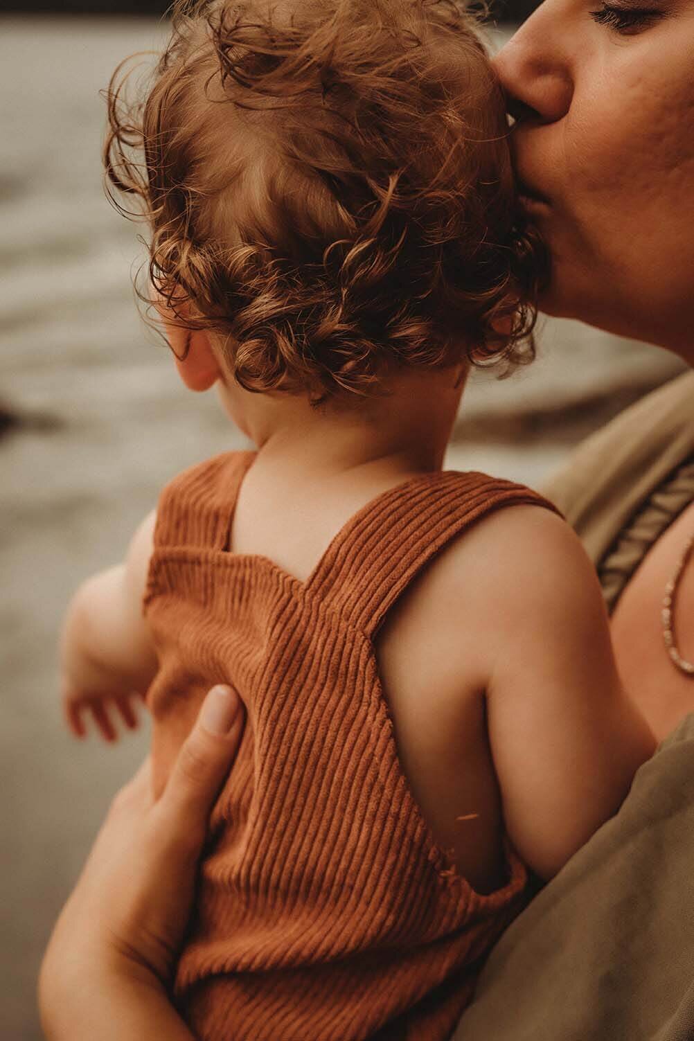 a mother holds her curly haired baby while she kisses the side of his head. he is facing away from the camera so we can see the details in his hair