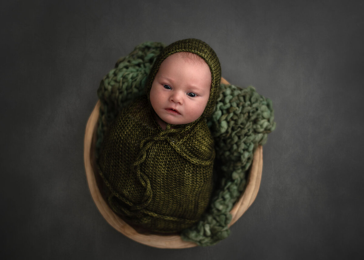 baby in a bowl with green wrap and bonnet looking up at camera by st. louis newborn photographer, Sutherland Photography