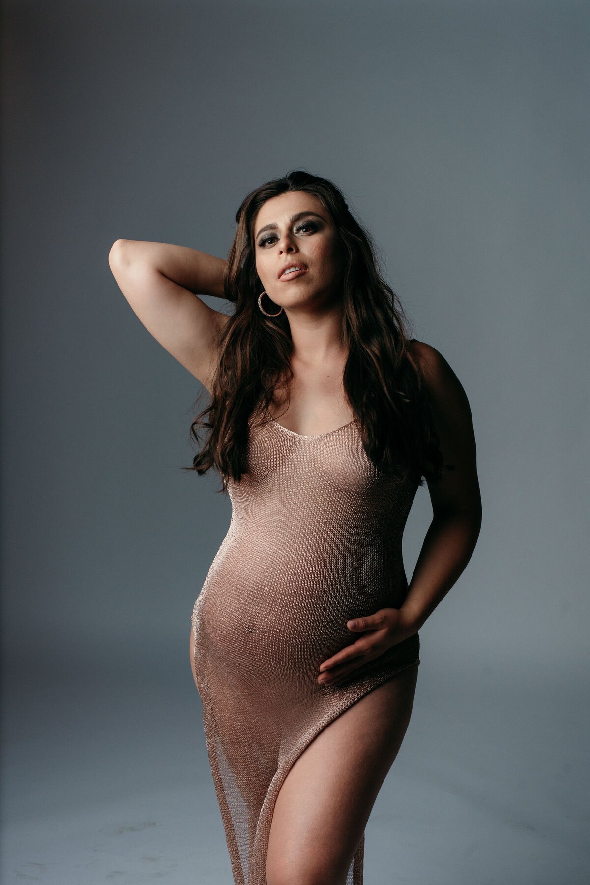 Pregnant woman wearing see through nude color knit dress with high slit holding tummy with one hand and other hand behind her head