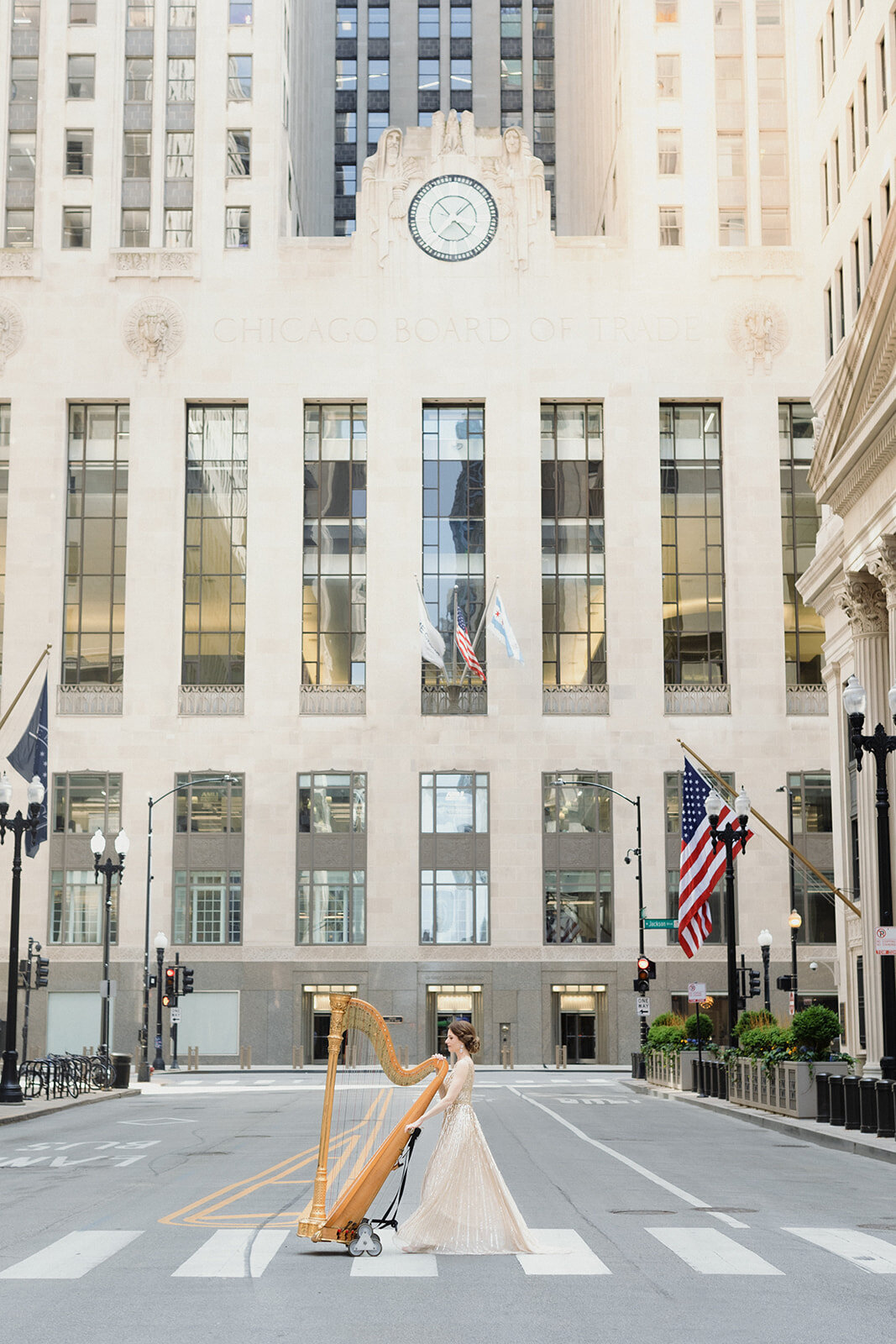 woman in pink gown is walking across crosswalk, pushing her harp. The crosswalk is in Chicago, with the Chicago Board of Trade in the background.