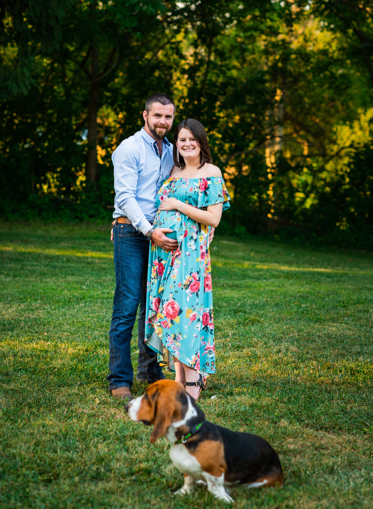 Expecting couple poses joyfully in a lush green park with their dog in the foreground