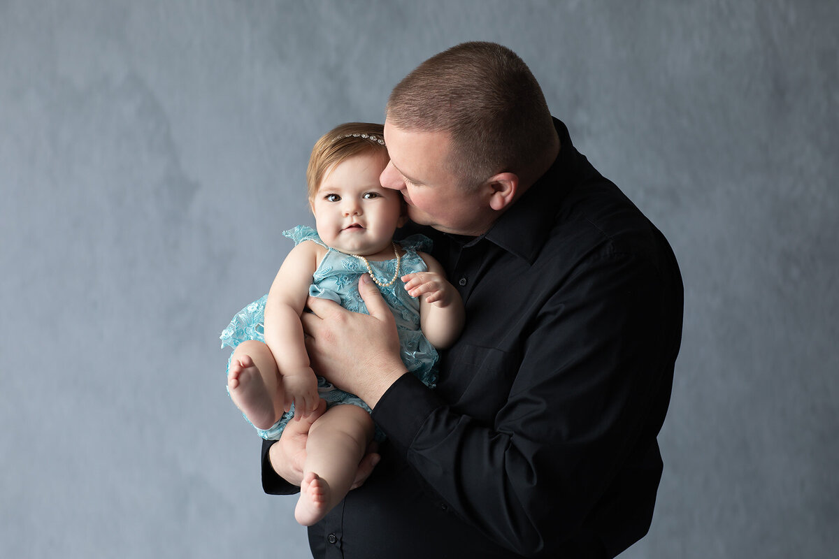 A dad kisses his baby girl on the cheek as he holds her close, both dressed in coordinating colors during a Dallas milestone photography session.