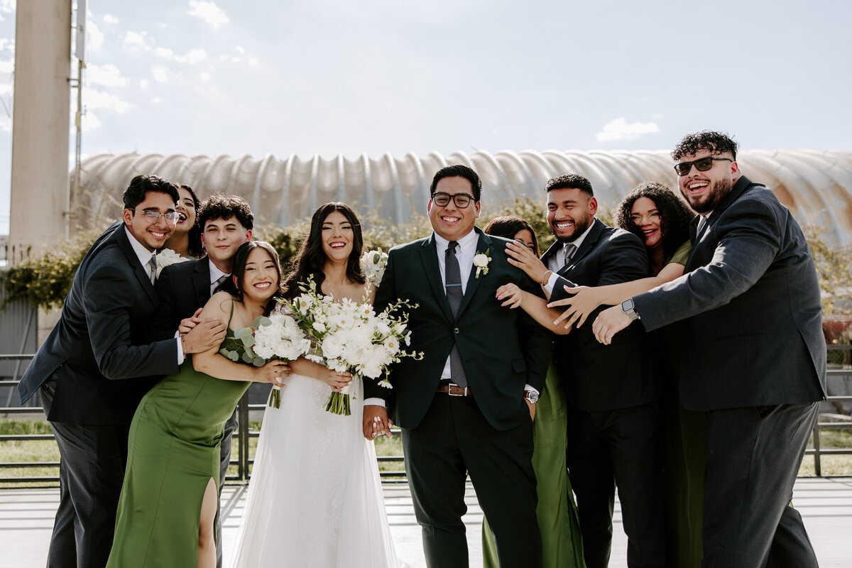 A couple smiles with their wedding party around them.