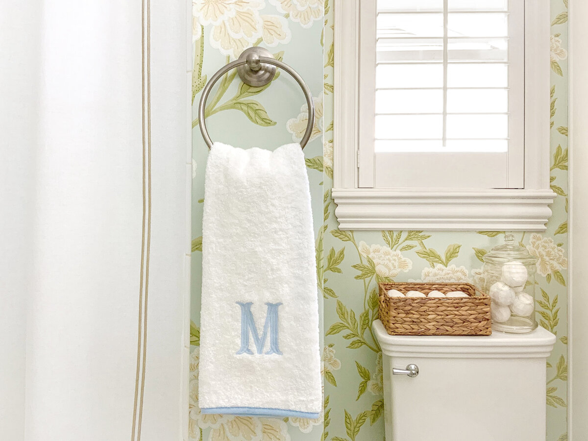 Floral wallpaper bathroom with white embroidered shower curtain and decoraitve hand towel with blue embroidered initial and toilet with woven basket and hand towels and bath bombs in glass canister
