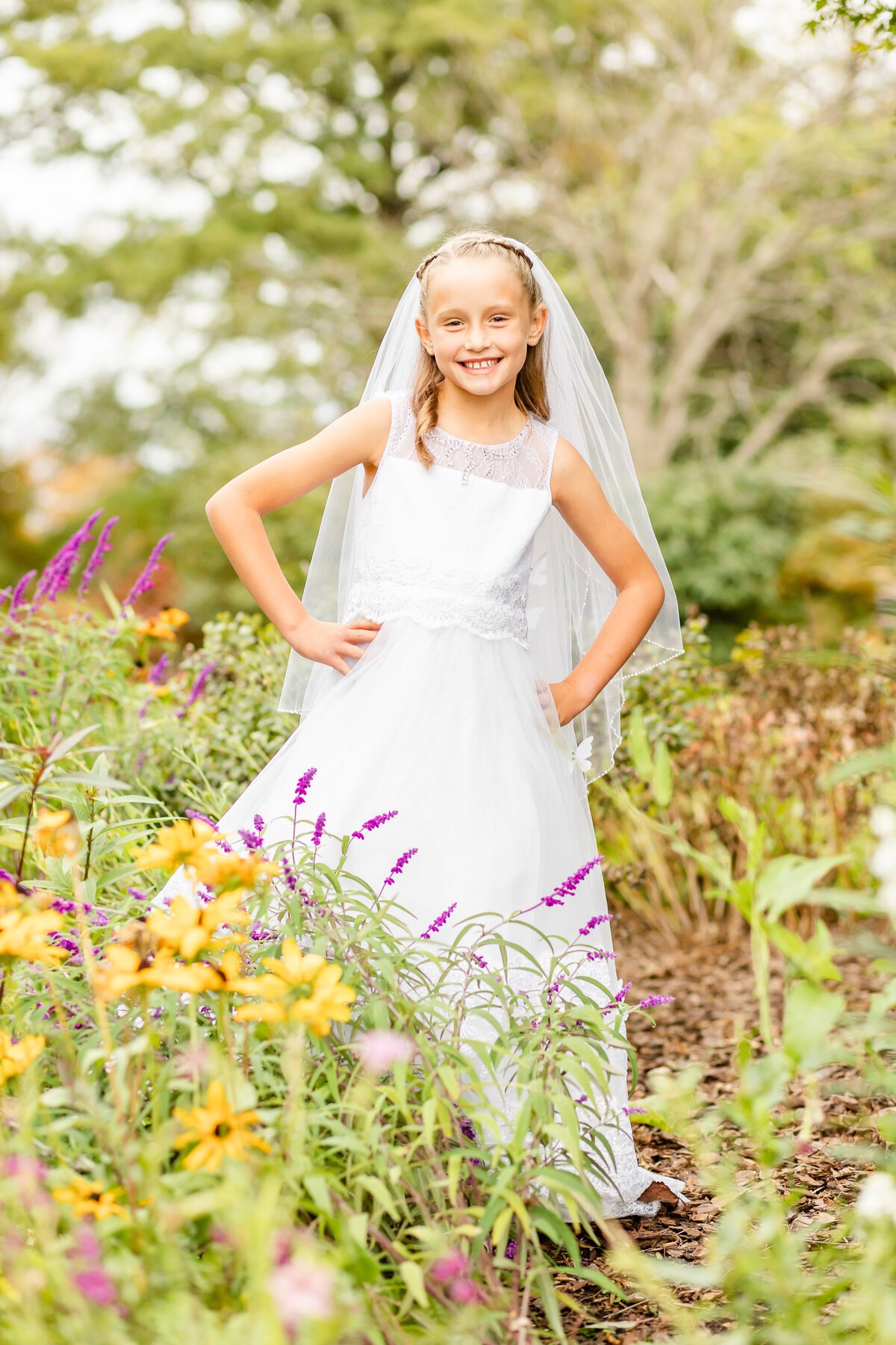 Girl's first communion standing in flowers with hands on hips smiling