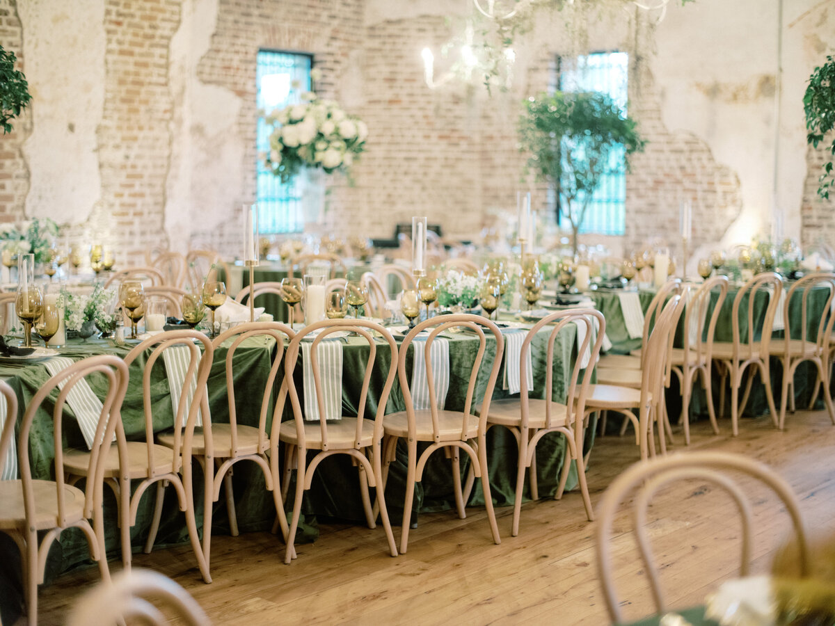 A beautifully styled long dining table featuring drapery in green tones, accented with golden glasses and delicate flowers, perfect for weddings or gatherings.