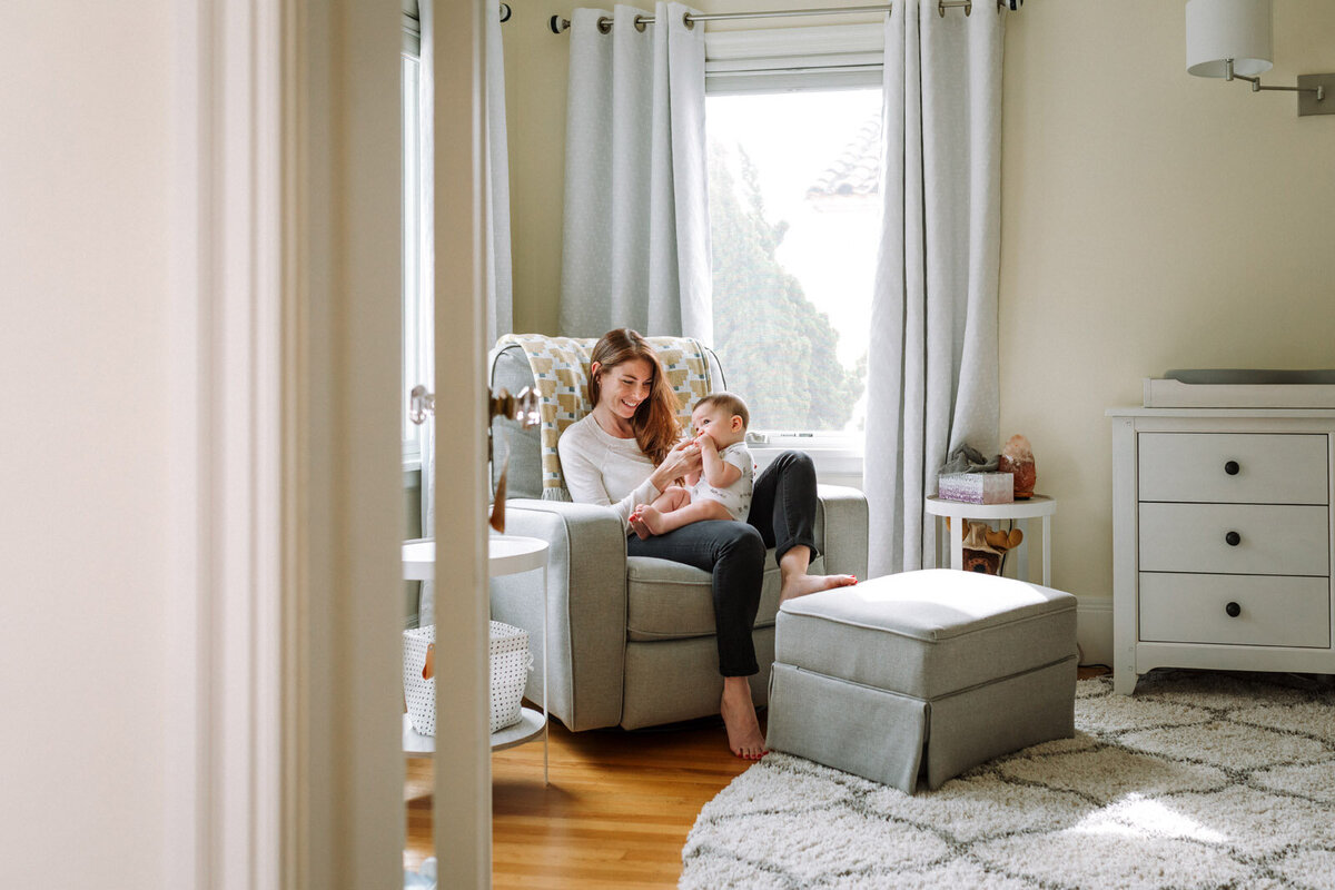 mom sitting in chair in nursery propping her foot on stool and holding baby in her arms smiling