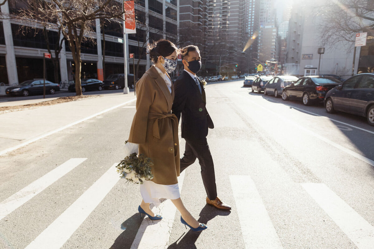 The bride, who is wearing a brown coat and holding her flower bouquet, and the groom, are crossing the streets of Brooklyn.