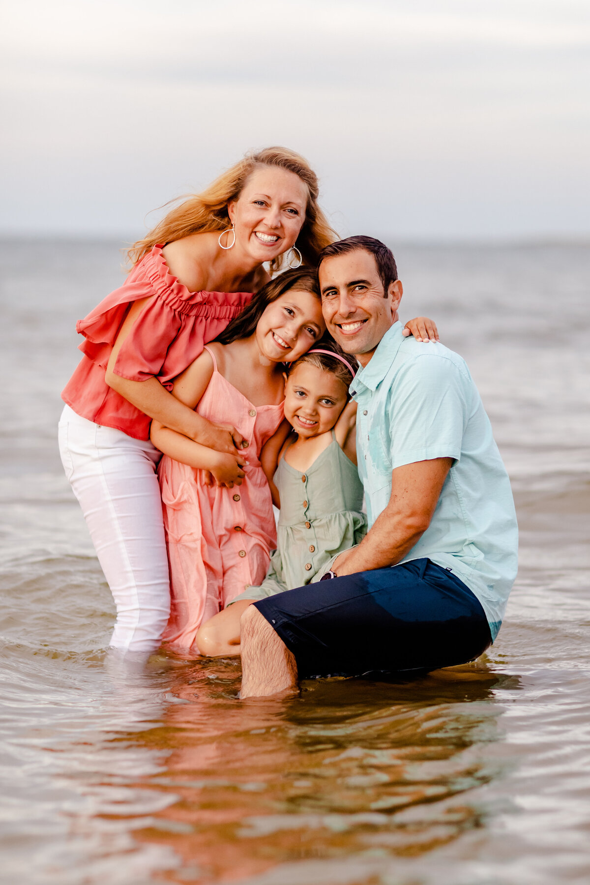 A joyful family of four dressed in soft pastel tones cuddles close in the gentle bay waves in Virginia Beach, their laughter and love radiating as sunset paints the sky.