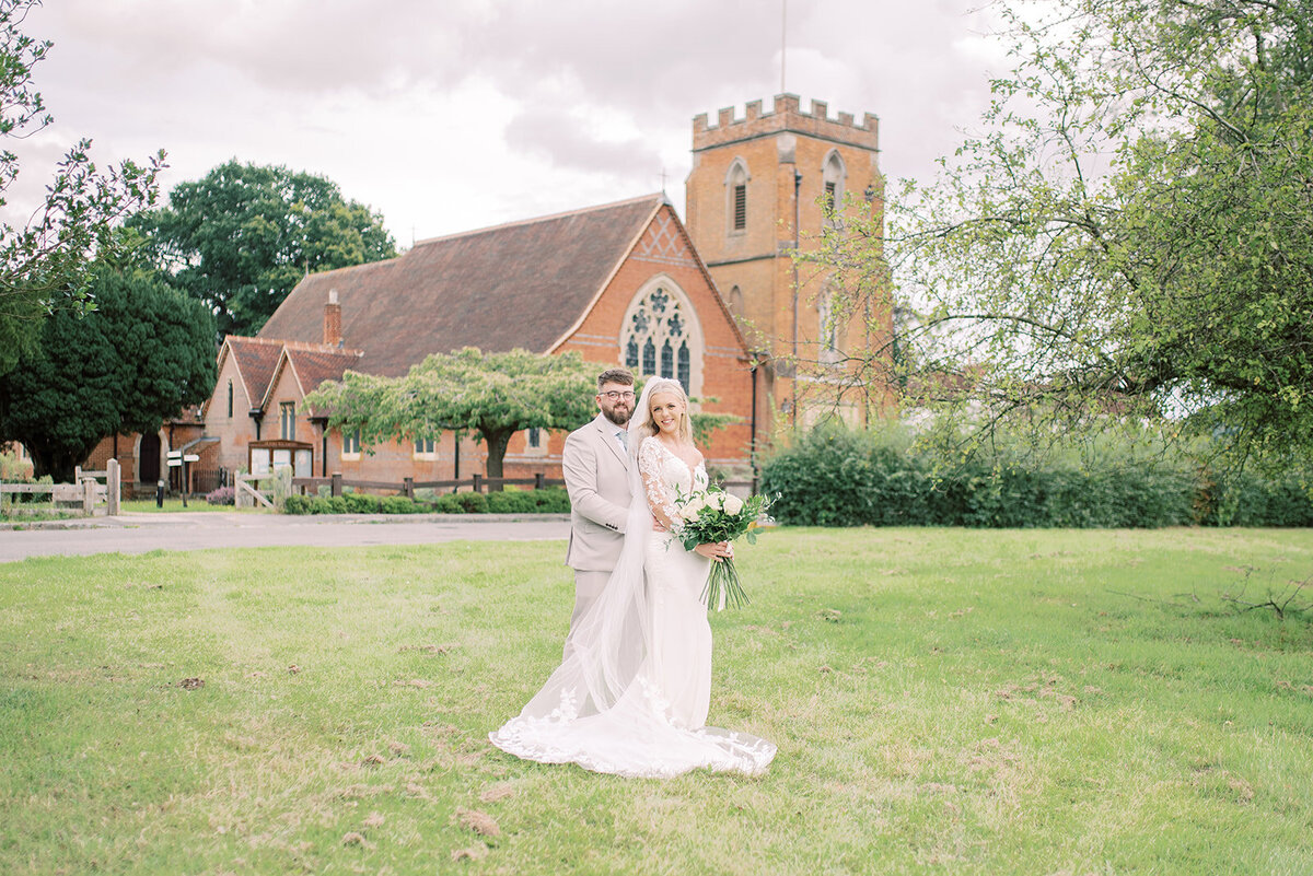 Bride and groom outside a church in surrey. The groom is stood behind the bride and has his hands around her waist. the bride is holding her bouquet at waist height and they are smiling at the camera