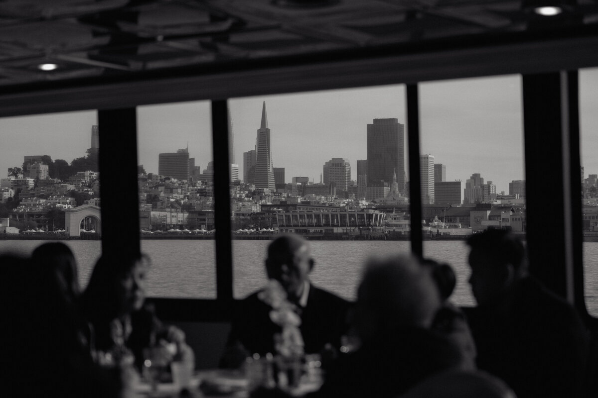 Guests dining onboard yacht with iconic San Francisco cityscape visible through windows, intimate documentary photography.