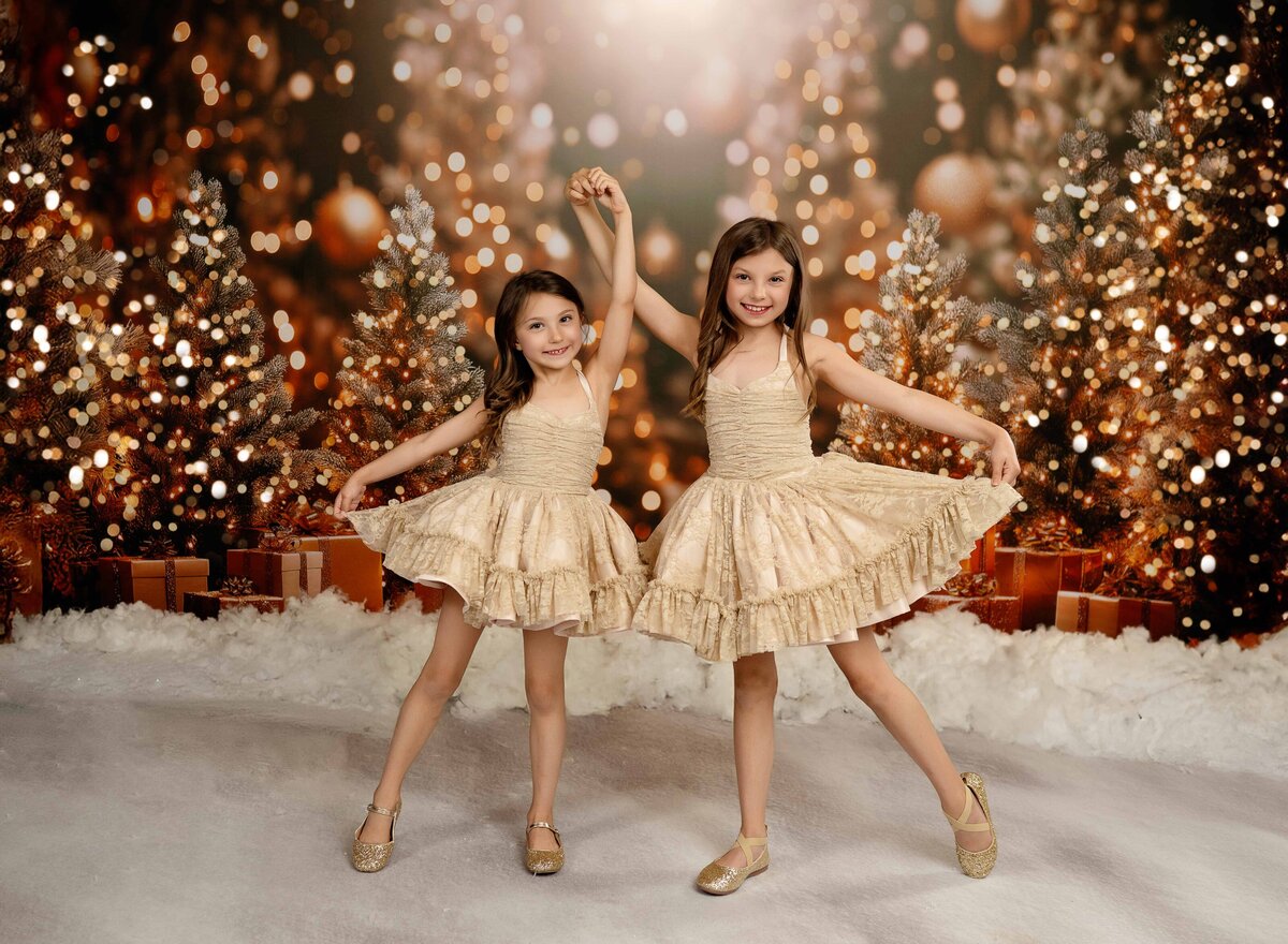 2 girls in gold dresses dancing and posing in front of a Christmas backdrop with golden lit trees and presents. 