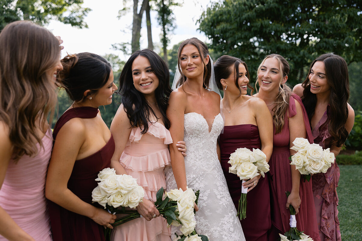 Bride with bridesmaids in romantic pink and burgundy dresses smiling together during an outdoor mountain wedding in Highlands, North Carolina.