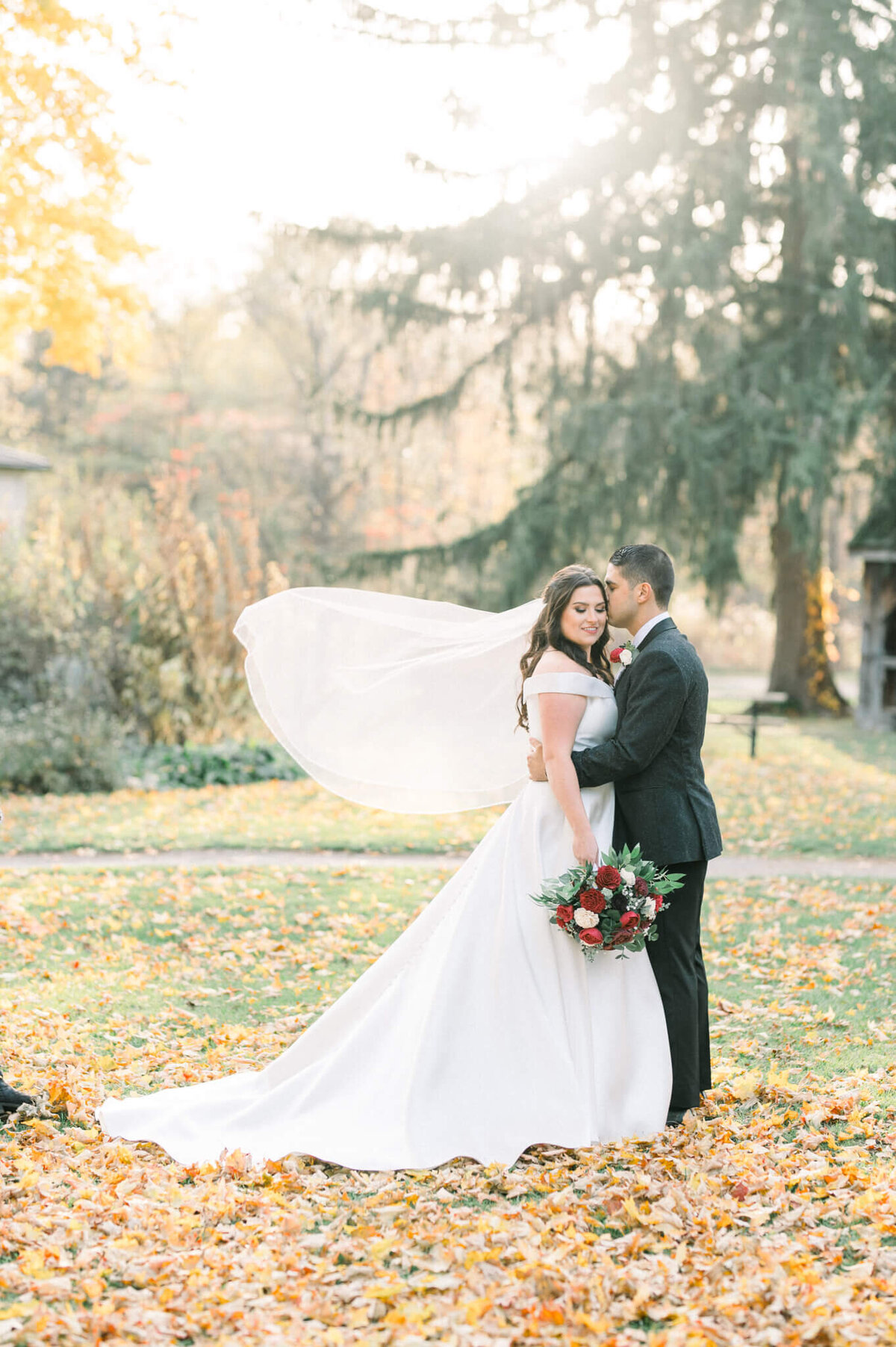 Bride and groom standing in the middle of fallen leaves as the wind blows  brides veil. Captured by Niagara wedding photographer Kristine Marie Photography