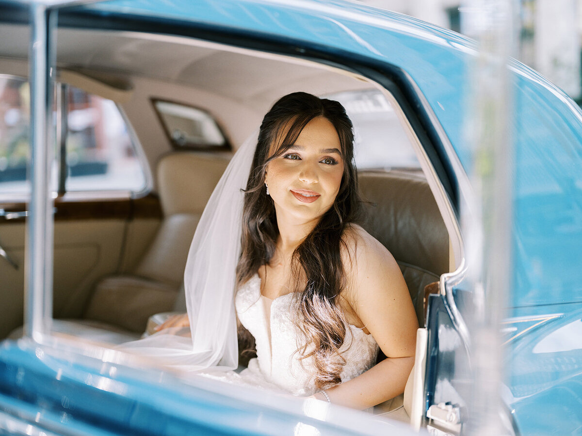 A bride with long, dark hair and a veil sits inside a vintage blue car, looking out the open window with a calm expression, ready for her Fairmont Palliser Wedding in Calgary.