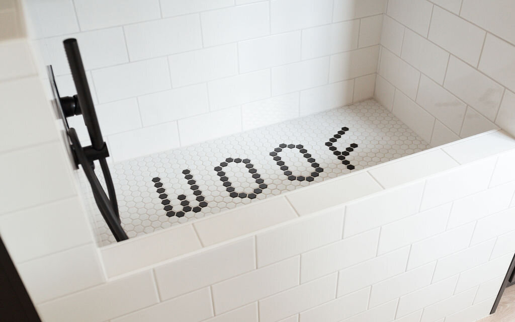 A close look at a modern dog wash room. The washing station is tiled with subway tile and the smaller tiles at the bottom of the tub read ‘woof’. The hardware on the dog shower is black.