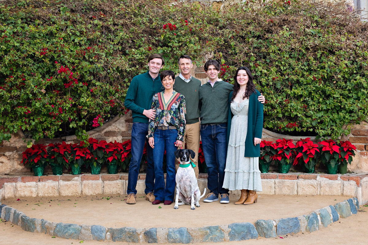 A family of five with their black and white dog standing in front of a wall of green plants and red flowers.
