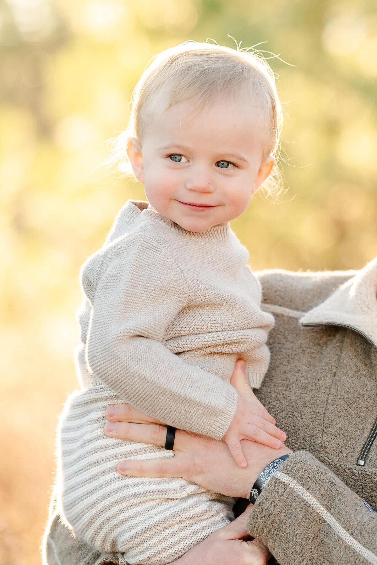 A toddler dressed in neutrals, is held by his dad. He looks off to the side with a smirk on his face.