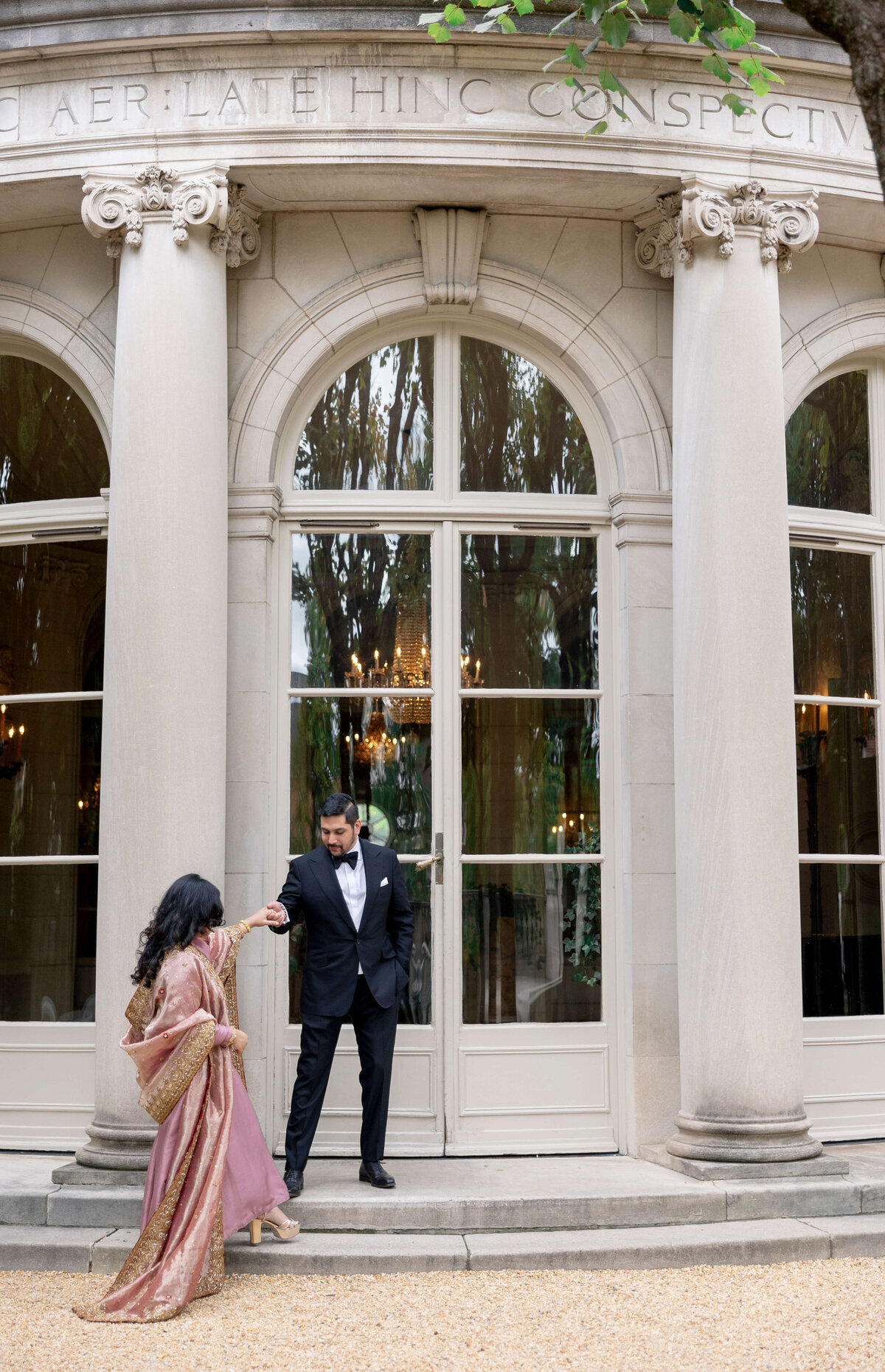 A man in a suit elegantly holds the hand of a woman in a pink sari as they walk beside a grand building with tall columns and large arched windows. Chandelier reflections can be seen in the windows.