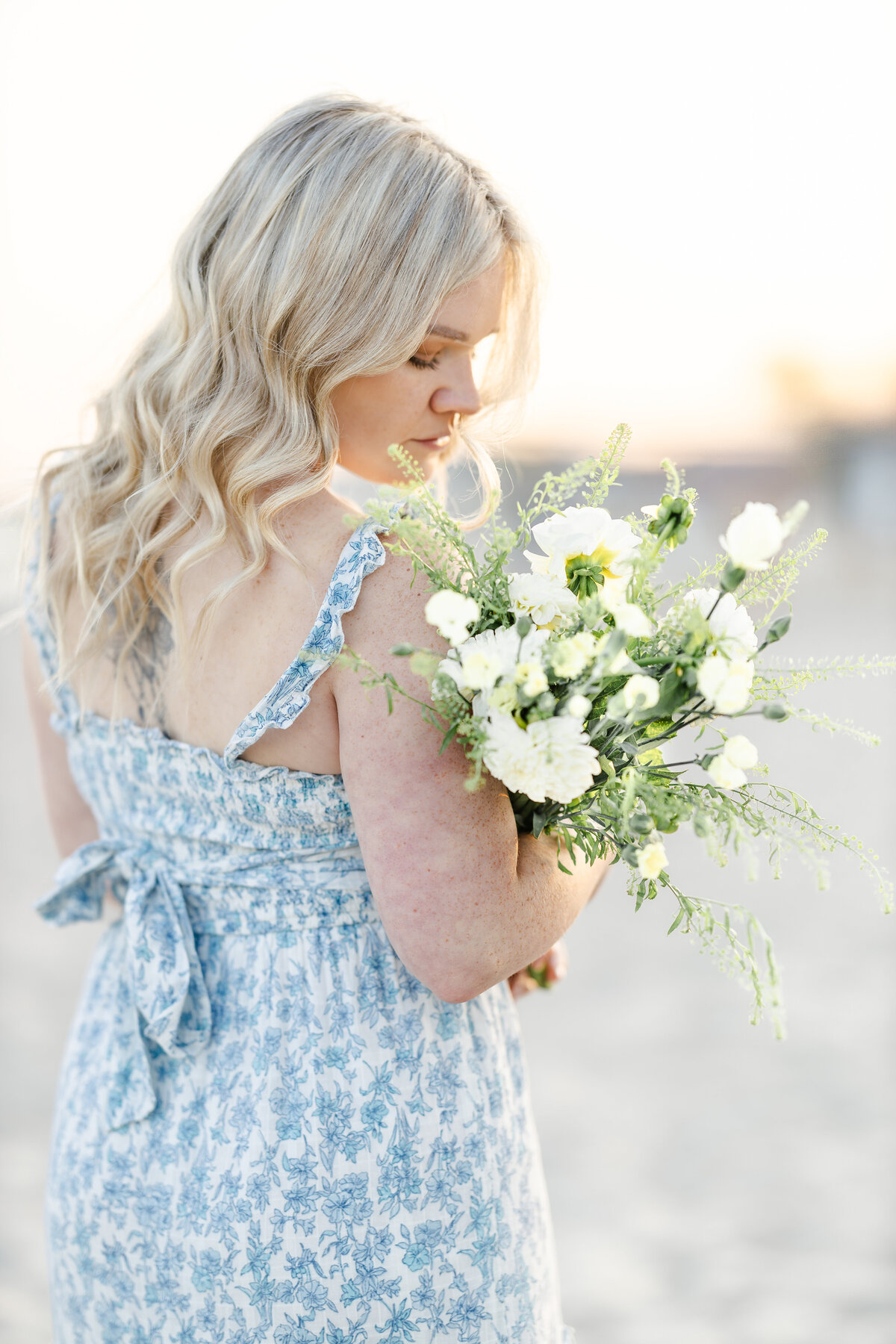 Young blonde woman in a floral blue dress walks along the beach, smelling a bouquet of flowers as the ocean breeze flows around her.