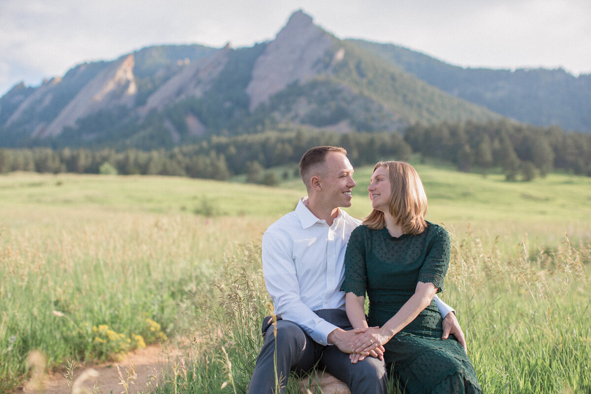 The engaged couple smile affectionately at each other while sitting on a rock in a field with an epic view of the Boulder flatirons