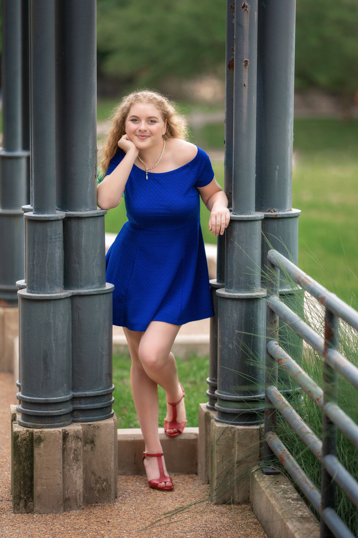 Girl in royal blue skater dress and red heels.  The girl is standing in between green metal poles on the Loyola campus.  She has one hand near her face and is resting her cheek on it.  She is smiling and looking at the camera.  She has long blonde curly hair.