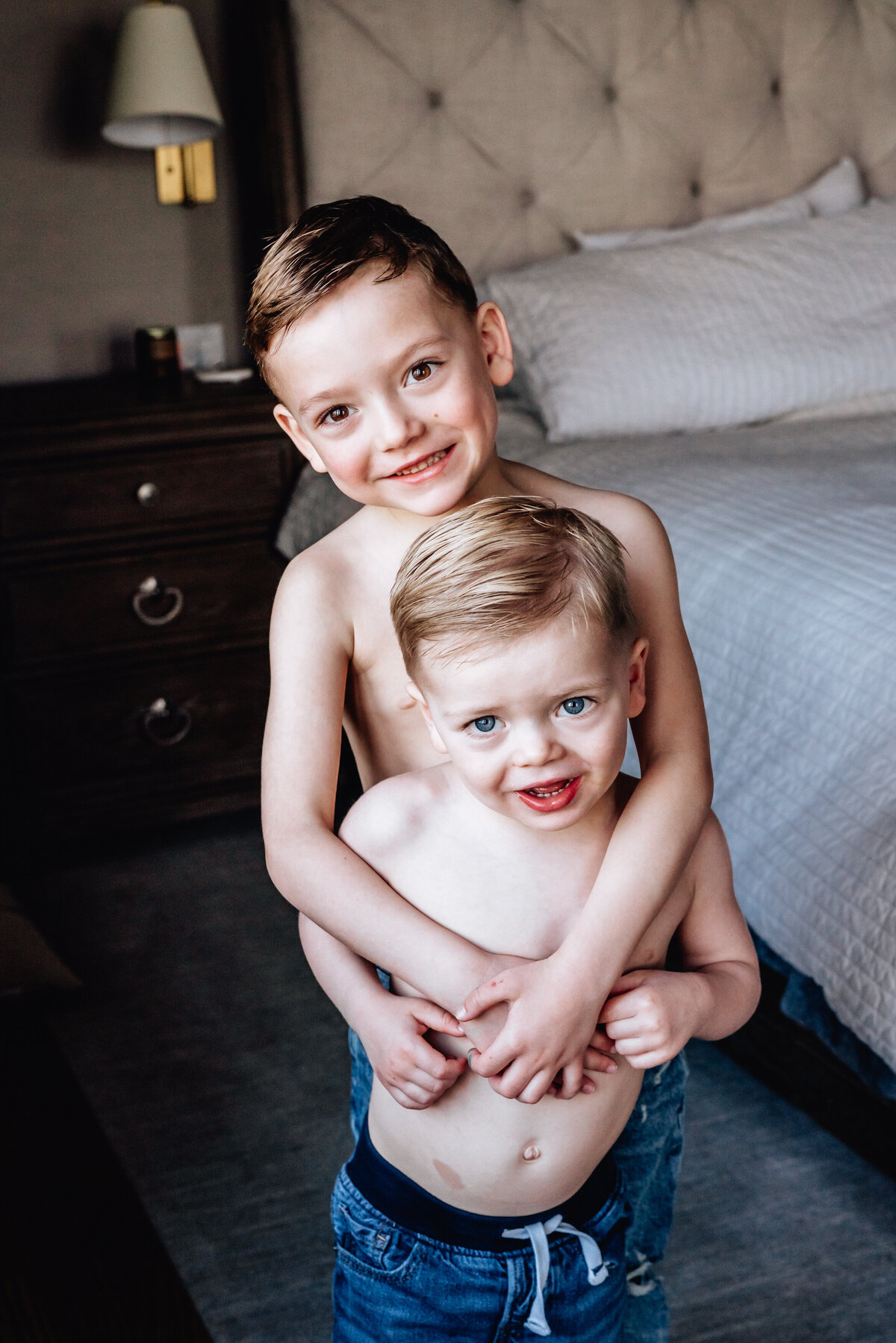 Two young boys, shirtless and smiling, stand in front of a bed. The older boy wraps his arms around the younger boy from behind, both looking happily at the camera.