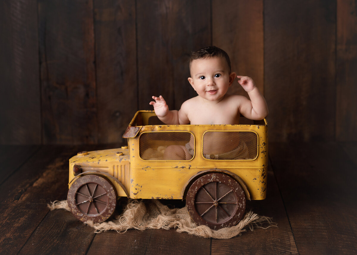 Baby Boy 6-month milestone photoshoot at West Palm Beach photography studio. Baby boy is sitting in a vintage inspired yellow metal truck smiling at the camera.