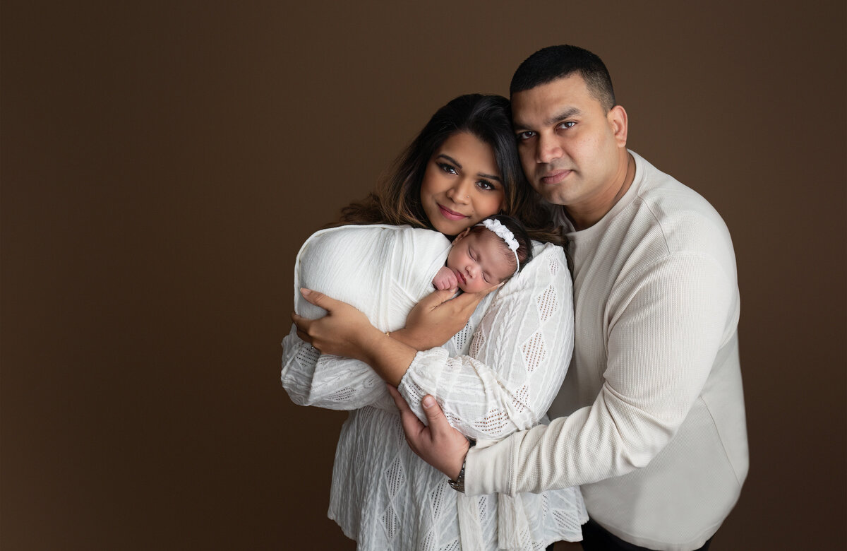 A classic family studio portrait featuring parents holding their newborn in front of a seamless mocha background. Captured at Meraki Photographic Creations in Paris, Ontario.