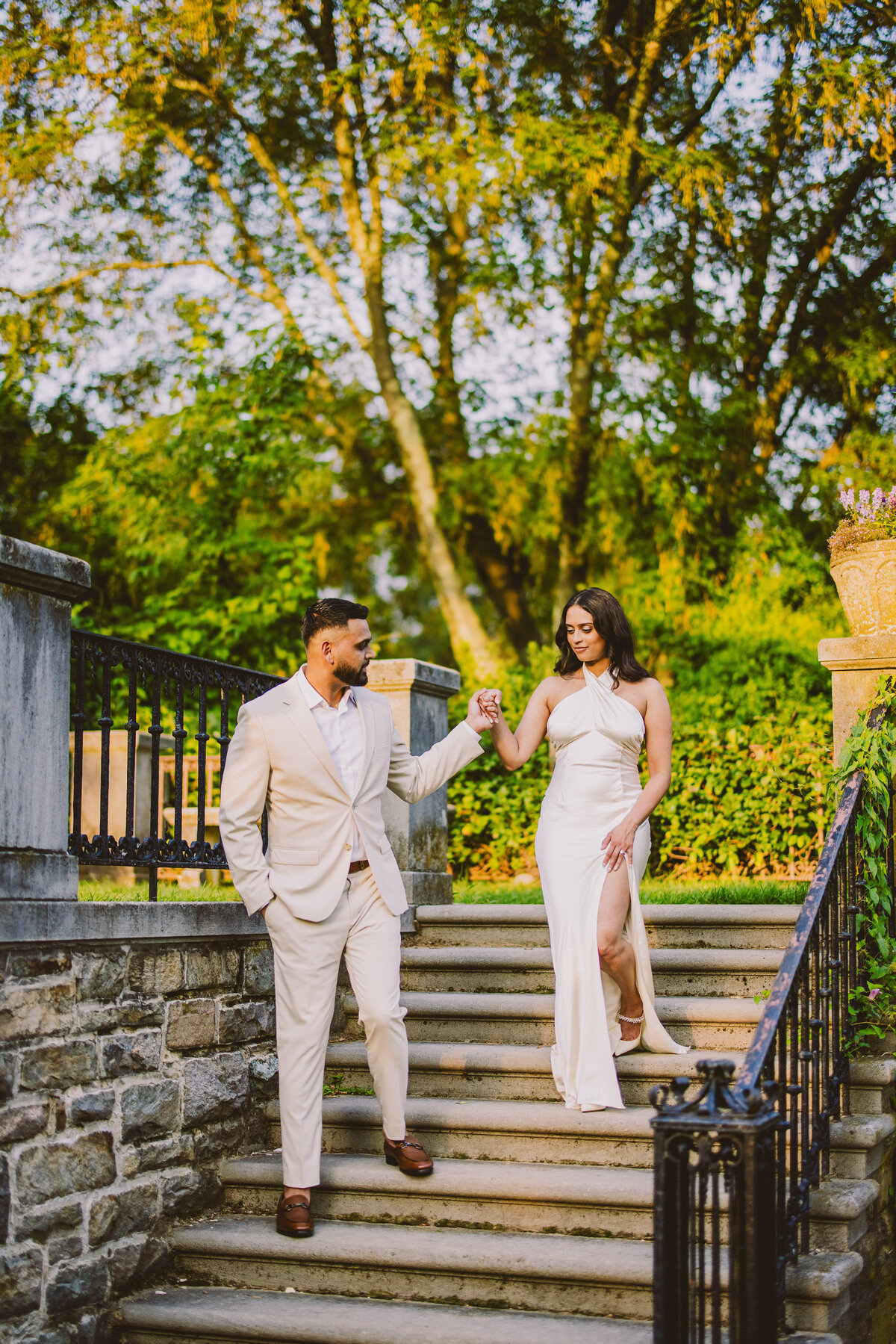 An engaged couple wearing white formal outfits walking down some steps at the nj botanical garden in ringwood state park