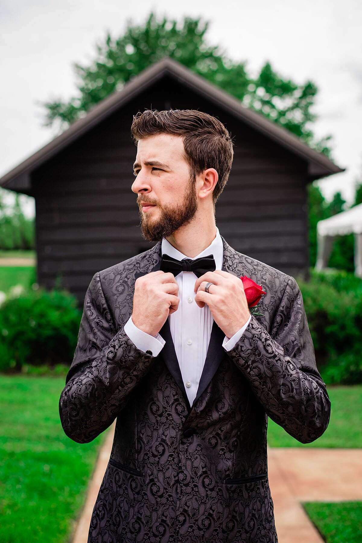 The groom, wearing a black silk tuxedo with an embroidered paisley pattern and red rose boutonniere adjusts his black bowtie as he stands in front of a rustic barn wood cabin at Arrington Vineyards.