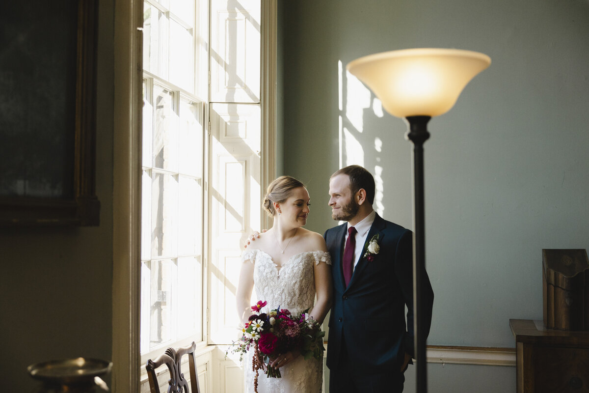 A newlywed couple standing by a window looking at each other and smiling
