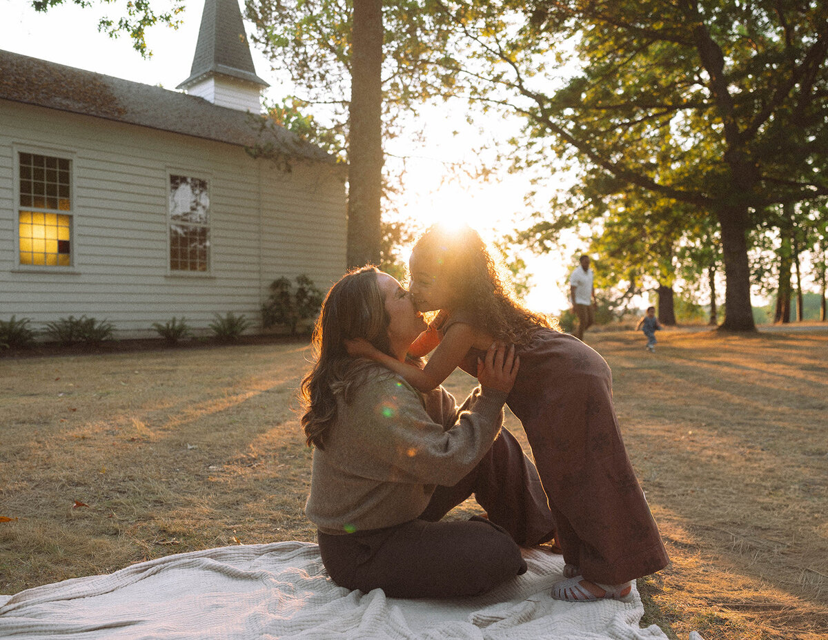Mother and daughter sharing a sweet kiss in front of a small chapel at sunset, part of a candid family session in Orange County.