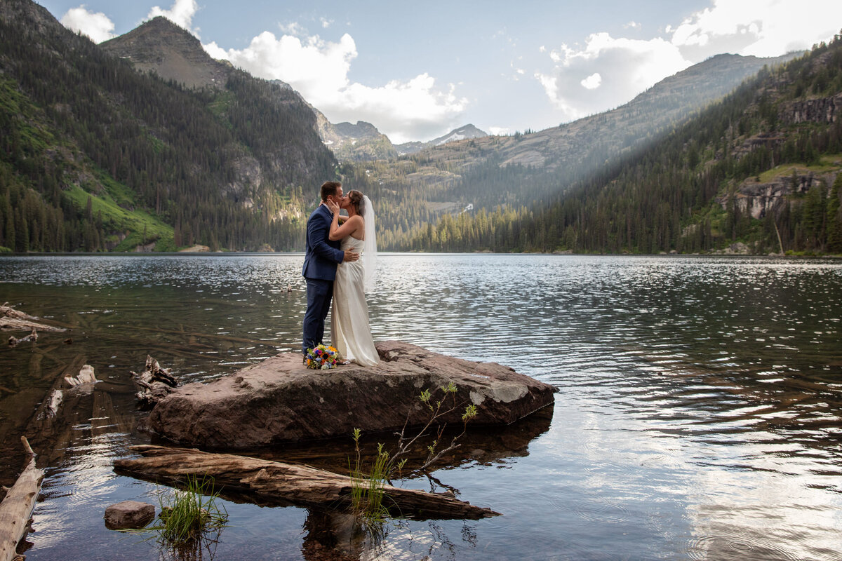 A bride and groom share a kiss on top of a rock in the middle of a lake in Montana.