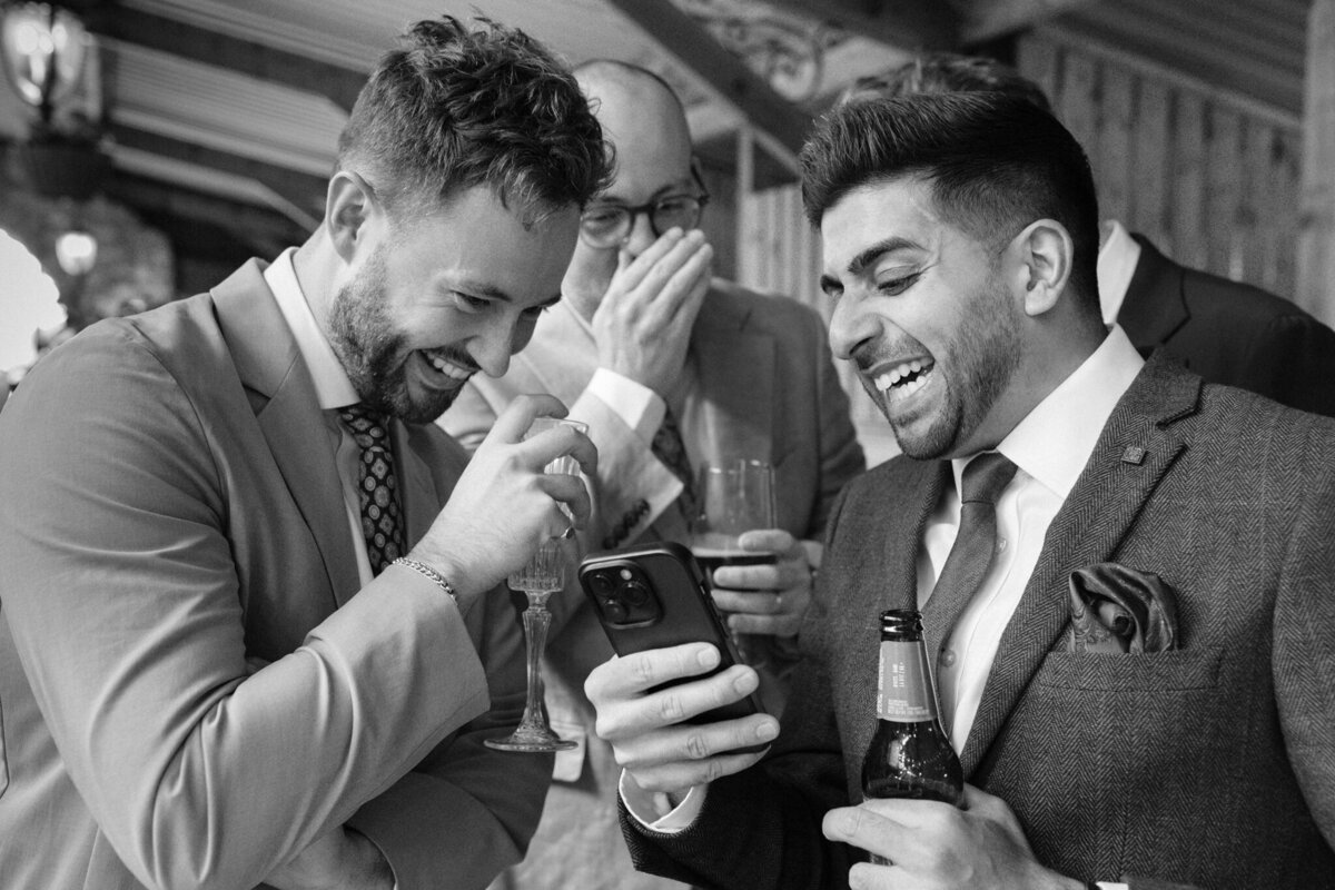 "A candid black-and-white moment from the wedding at Willow Marsh Farm, where a group of groomsmen share a laugh. Two men, one holding a drink and the other a phone, are focused on something on the screen, both grinning widely. In the background, a third man laughs with his hand over his mouth, creating a lighthearted, joyful scene filled with camaraderie and celebration.