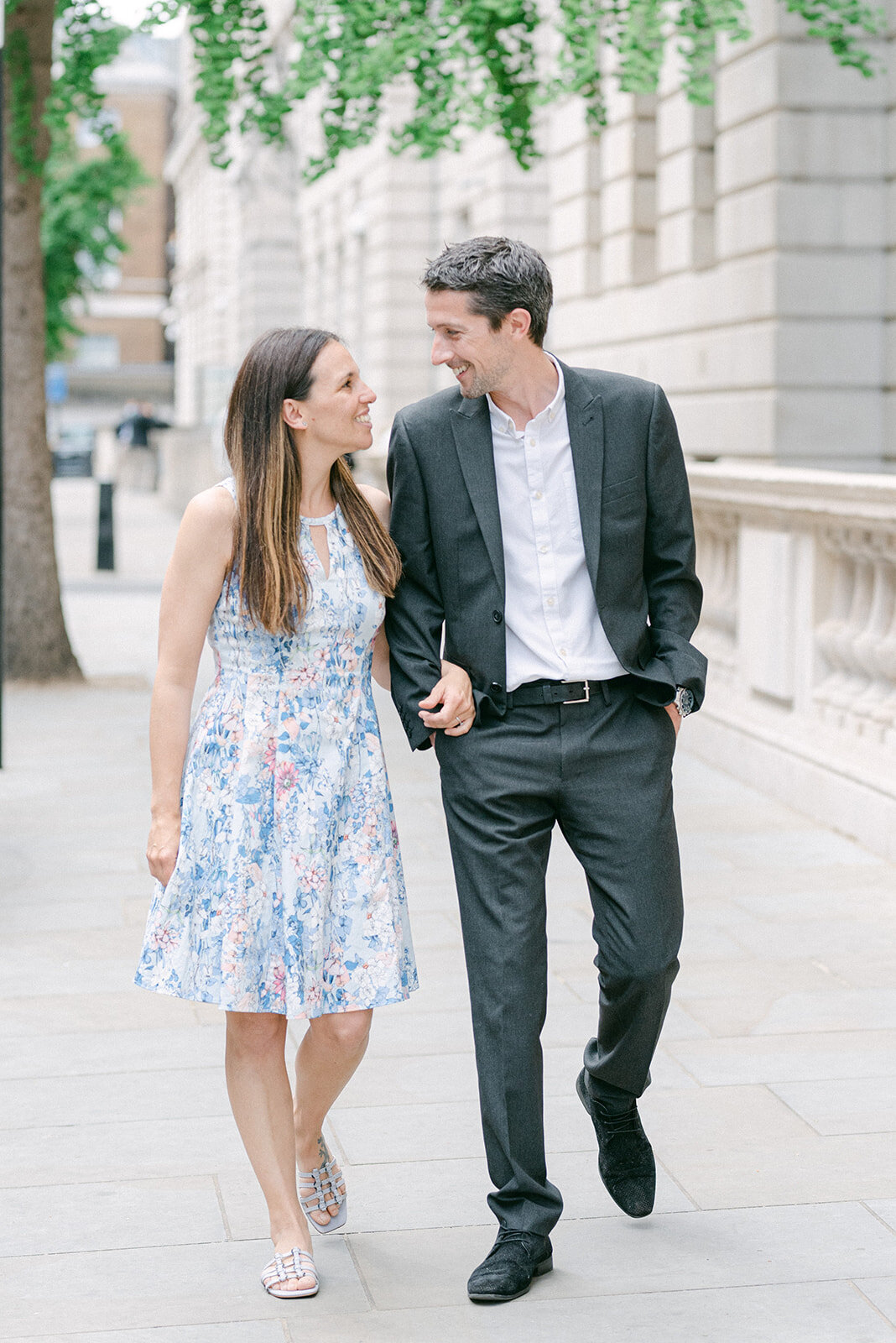 Couple strolling through London for an engagement photoshoot, The man is in a black suit and the lady is wearing a blue flowery dress and they are looking at each other smiling